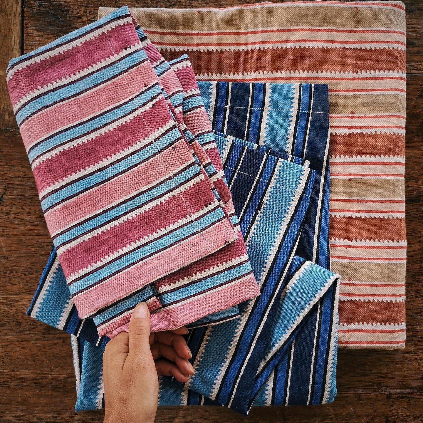 Overhead image of stacked folded fabrics in brightly colored stripes on a rustic wooden table with a womans hand holding the top fabric. 