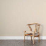 A wooden chair stands in front of a wall papered in a dotted diamond grid in white on a cream field.