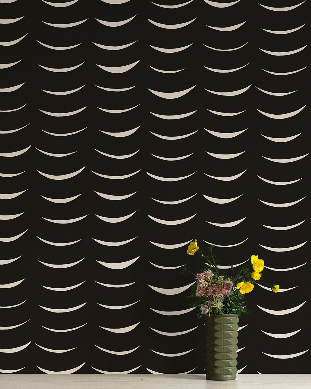 A vase of flowers stands in front of a wall papered in an abstract crescent print in cream on a black field.