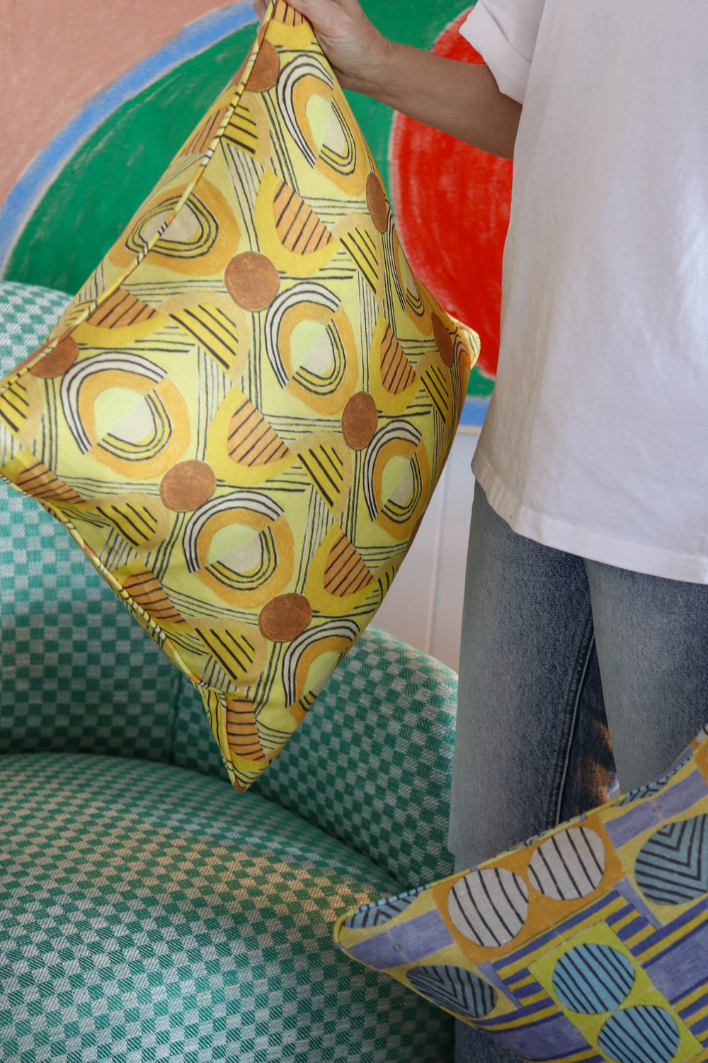 Woman in white tee shirt and jeans standing holding printed geometric pillows against a green checkerboard patterned arm chair.
