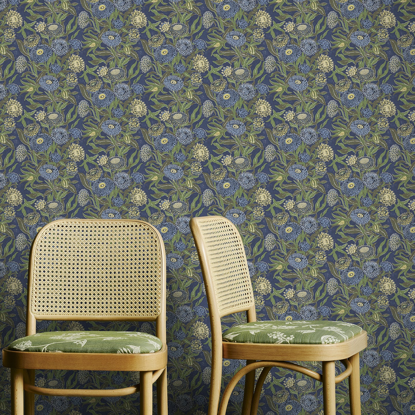 Two wooden chairs with floral cushions against a green and blue floral-patterned wallpaper