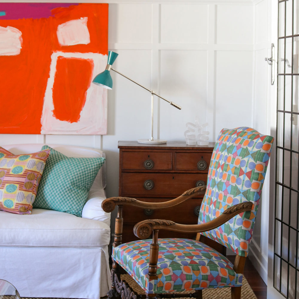 Bright living room setting with white paneled walls, bold red abstract paining, wooden chest, white couch with colorful geometric pillows, ornate wooden side chair with geometric upholstery and a natural fibre rug. 