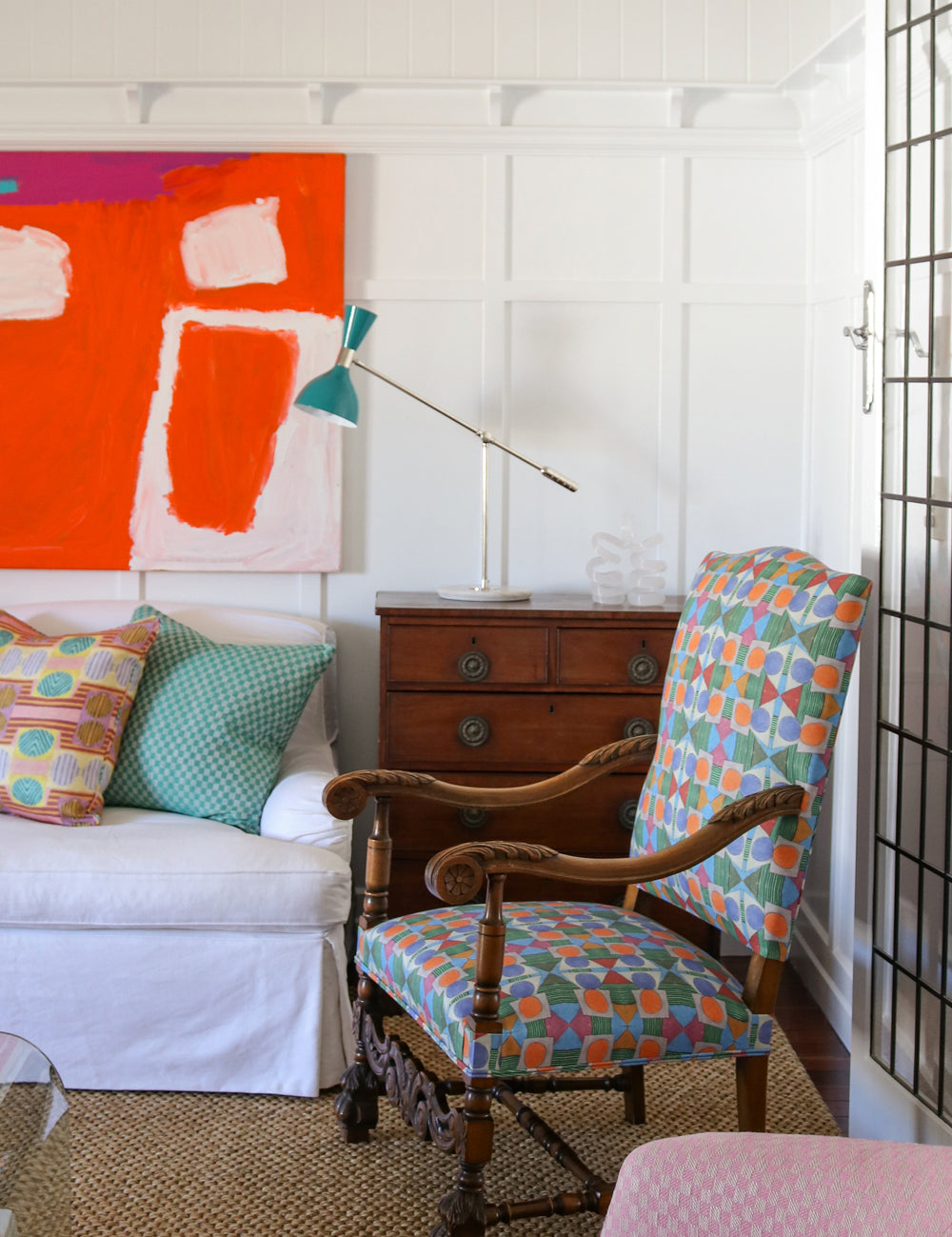 Bright living room setting with white paneled walls, bold red abstract paining, wooden chest, white couch with colorful geometric pillows, ornate wooden side chair with geometric upholstery and a natural fibre rug. 