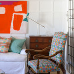 Bright living room setting with white paneled walls, bold red abstract paining, wooden chest, white couch with colorful geometric pillows, ornate wooden side chair with geometric upholstery and a natural fibre rug. 