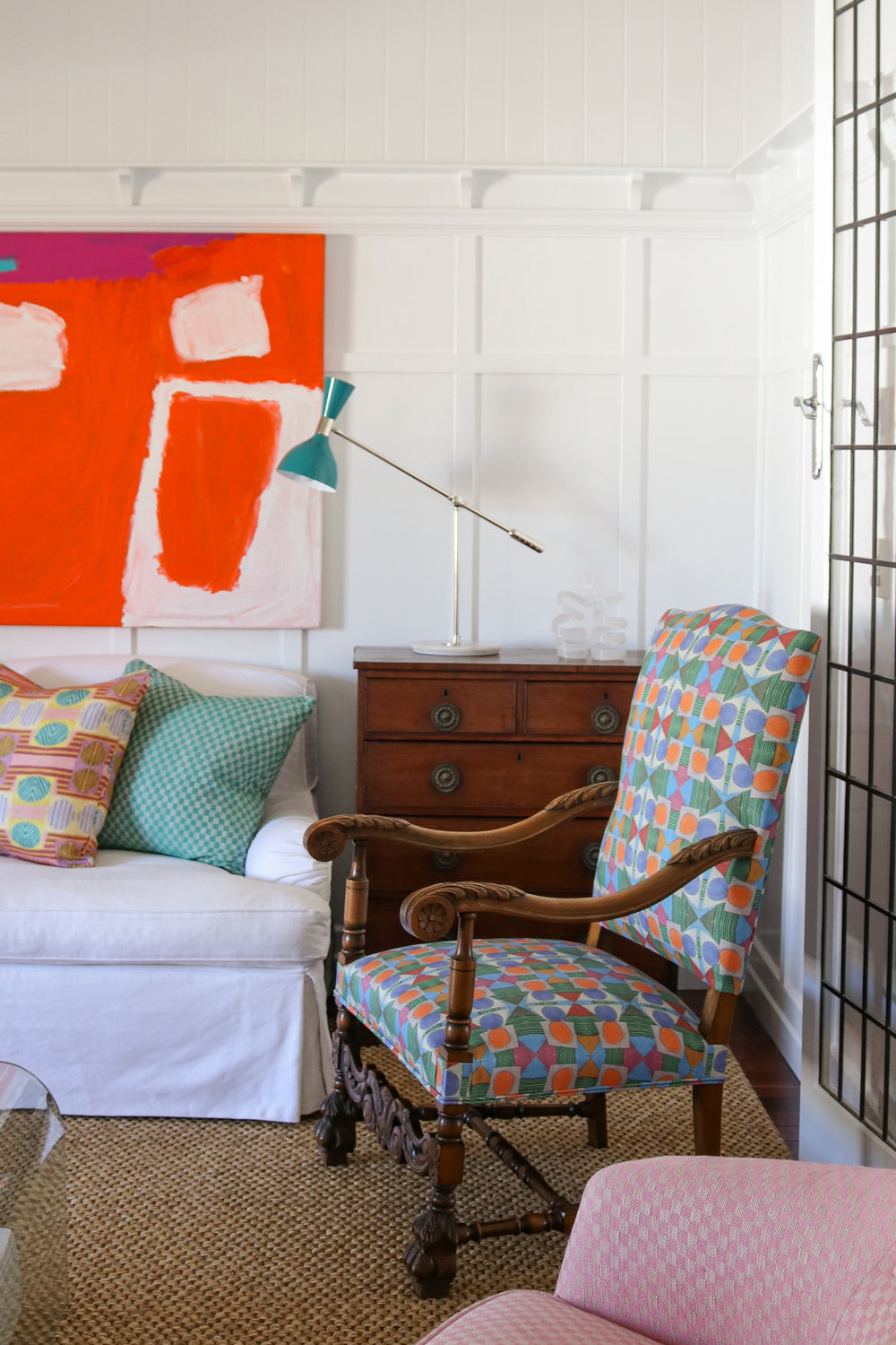 Bright living room setting with white paneled walls, bold red abstract paining, wooden chest, white couch with colorful geometric pillows, ornate wooden side chair with geometric upholstery and a natural fibre rug. 