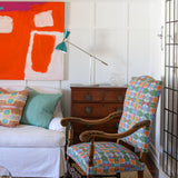 Bright living room setting with white paneled walls, bold red abstract paining, wooden chest, white couch with colorful geometric pillows, ornate wooden side chair with geometric upholstery and a natural fibre rug. 