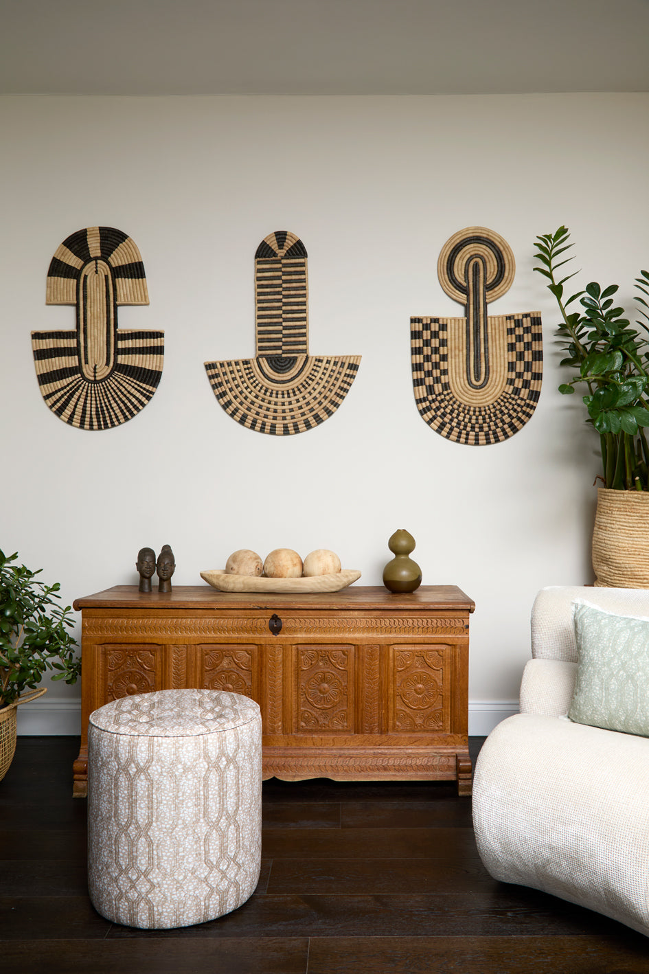 Living room scene with three black and tan sculptural woven panels, ornately carved wooden chest, baskets with house plans, white upholstered side chair and a stripe patterned footstool in beige. 