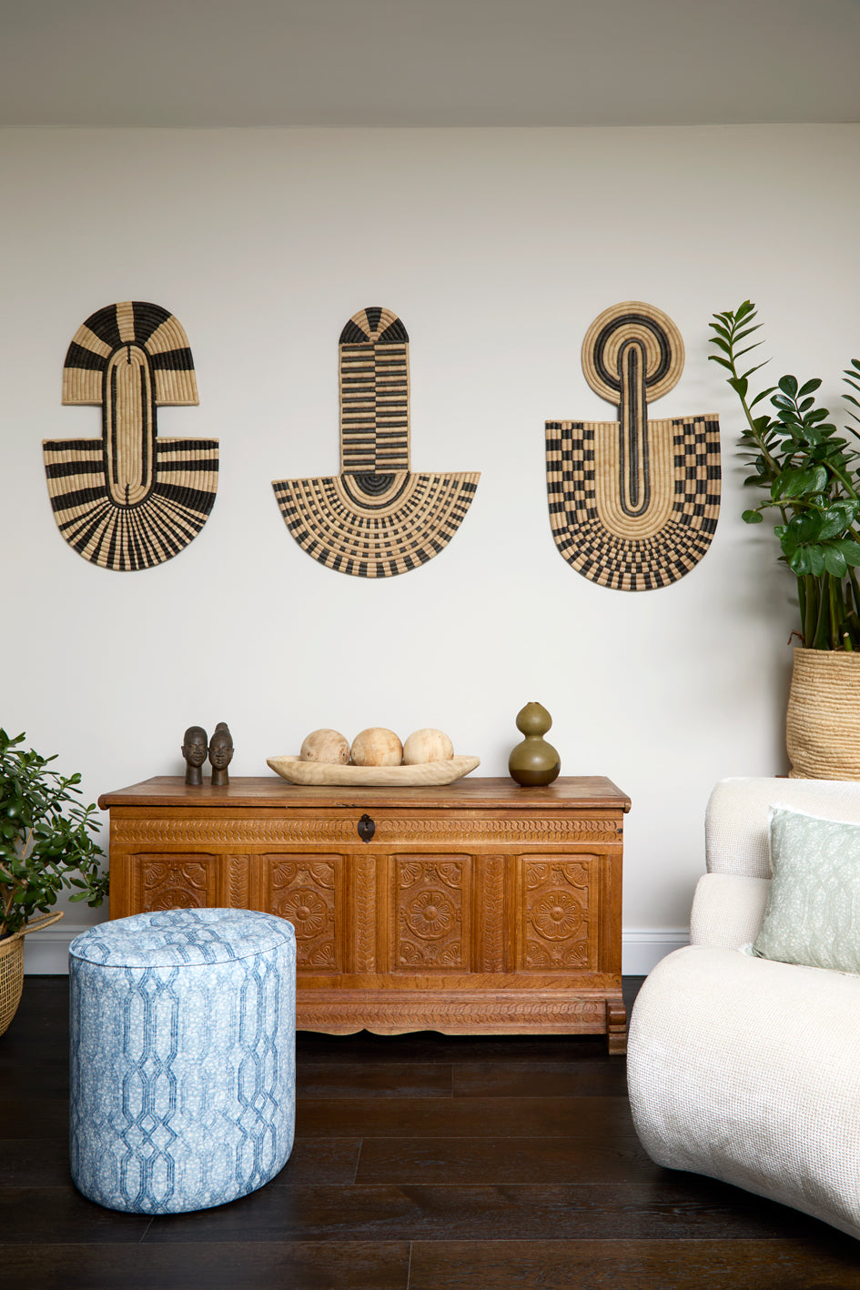 Living room scene with three black and tan sculptural woven panels, ornately carved wooden chest, baskets with house plans, white upholstered side chair and a stripe patterned footstool in blue.