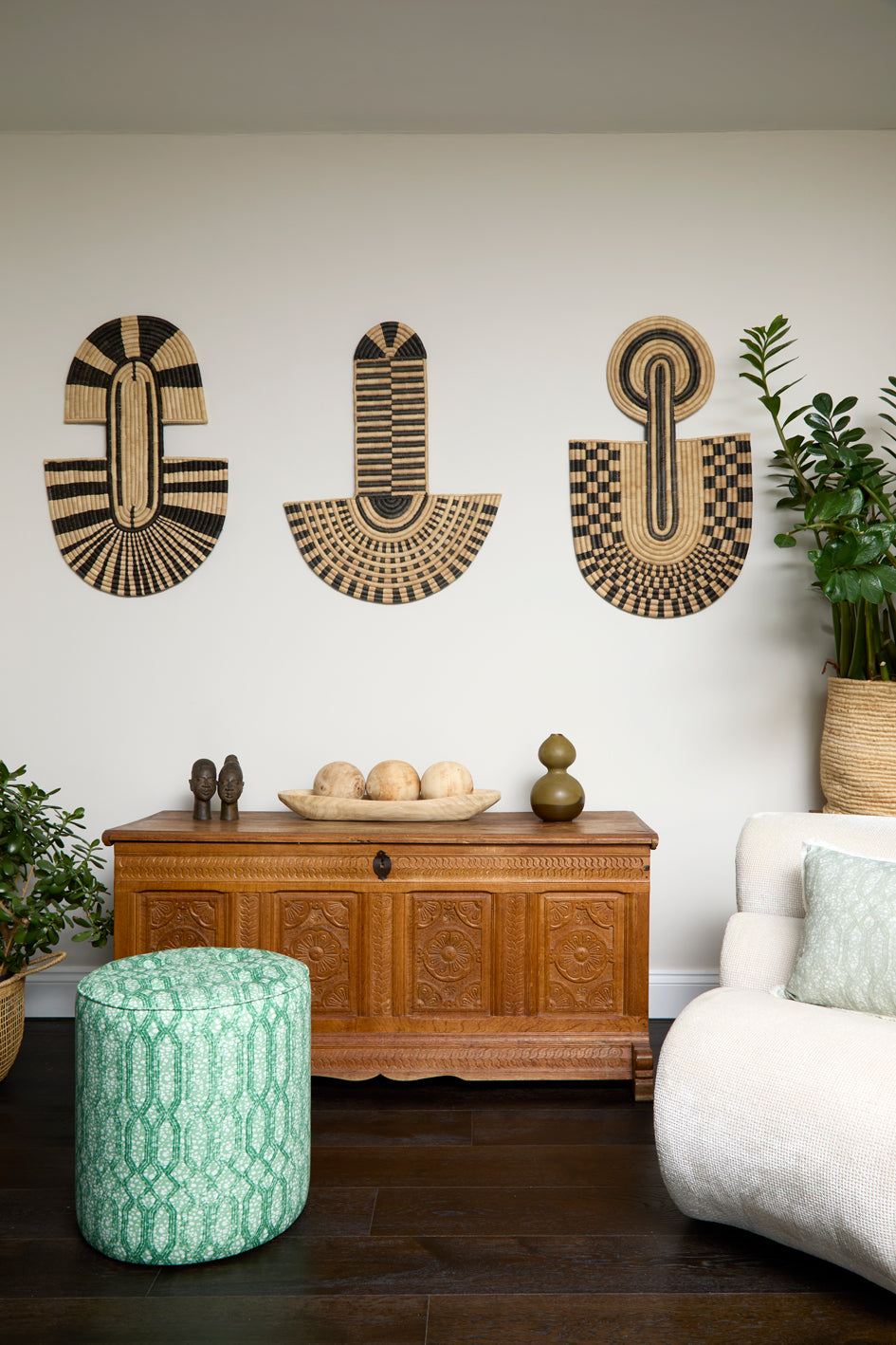 Living room scene with three black and tan sculptural woven panels, ornately carved wooden chest, baskets with house plans, white upholstered side chair and a stripe patterned footstool in green.