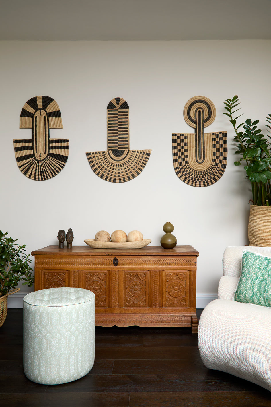 Living room scene with three black and tan sculptural woven panels, ornately carved wooden chest, baskets with house plans, white upholstered side chair and a stripe patterned footstool in pale grey.