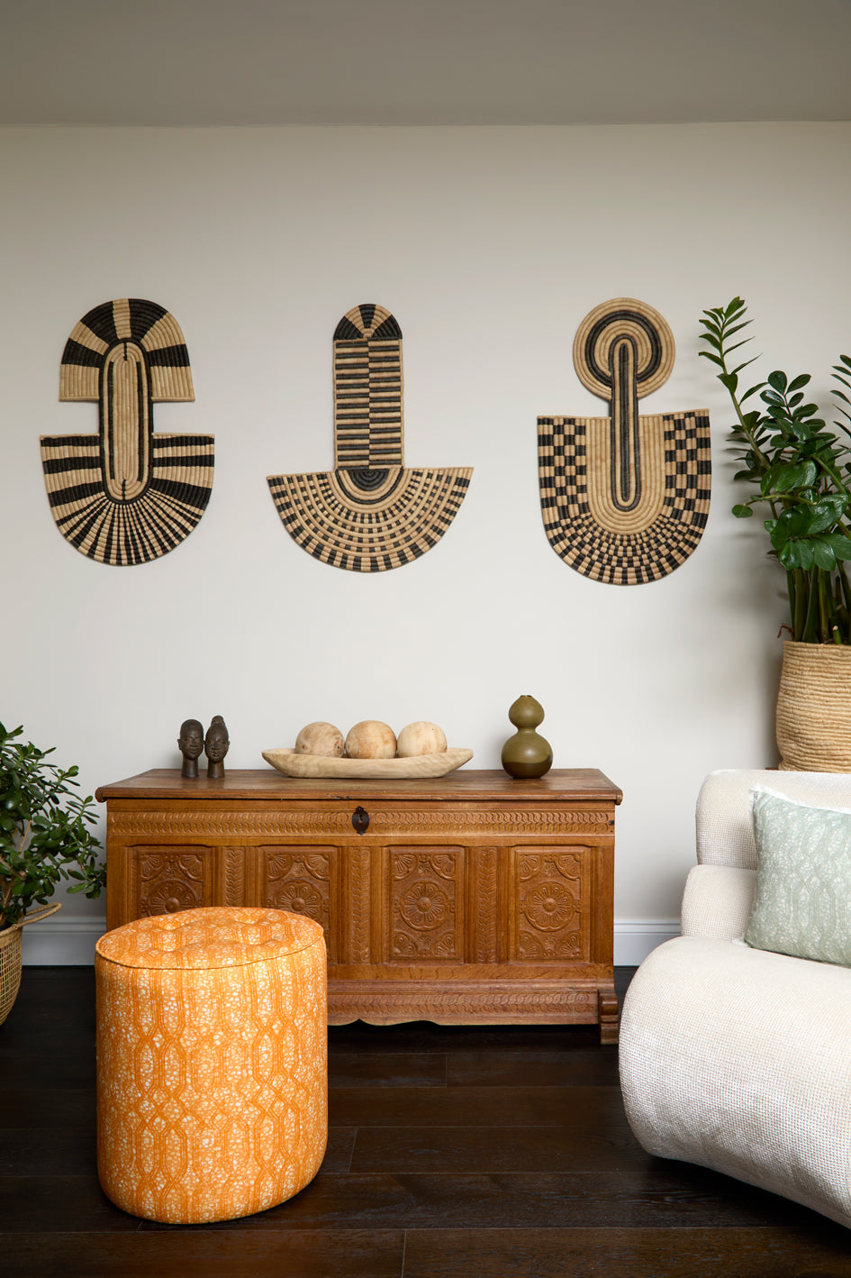 Living room scene with three black and tan sculptural woven panels, ornately carved wooden chest, baskets with house plans, white upholstered side chair and a stripe patterned footstool in orange.
