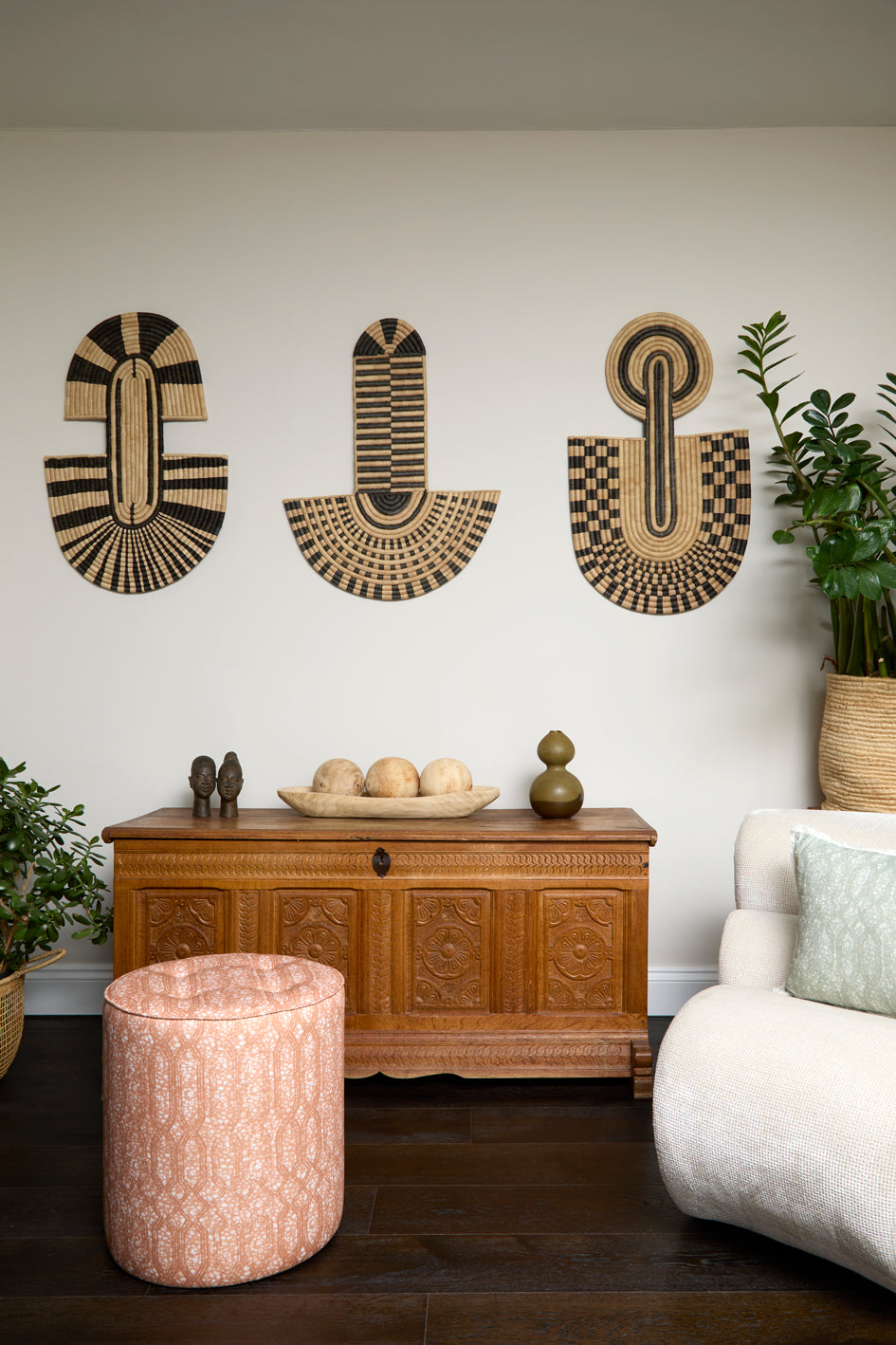 Living room scene with three black and tan sculptural woven panels, ornately carved wooden chest, baskets with house plans, white upholstered side chair and a stripe patterned footstool in orange. 