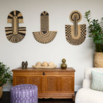 Living room scene with three black and tan sculptural woven panels, ornately carved wooden chest, baskets with house plans, white upholstered side chair and a stripe patterned footstool in purple.