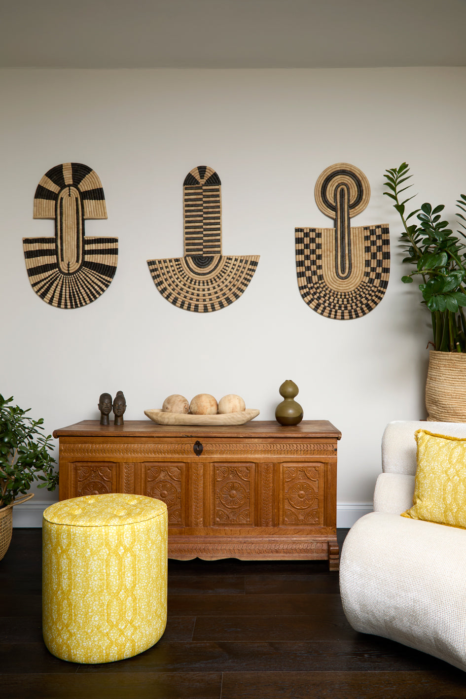 Living room scene with three black and tan sculptural woven panels, ornately carved wooden chest, baskets with house plans, white upholstered side chair and a stripe patterned footstool in yellow.