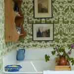 Bathtub with green patterned wallpaper, wooden shelf, and various items on the ledge.