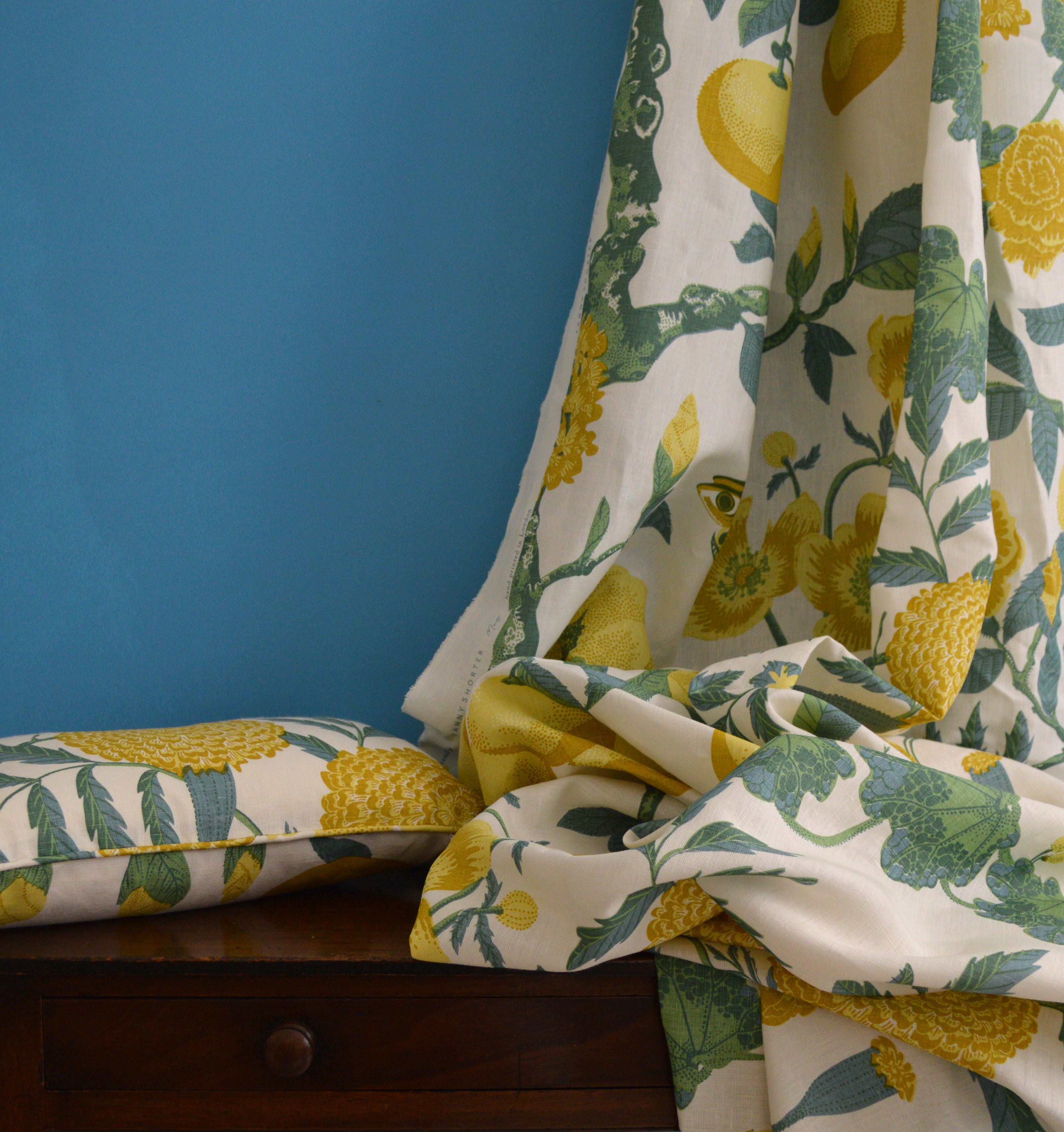 Floral and lemon patterned pillow and fabric draped over a wooden dresser with a blue background.