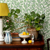 Wooden sideboard with plants, ceramic vesses, lamp and books against a green leaf wallpaper.