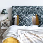 A bed with a headboard and pillows upholstered in a dense floral print in white, tan and green on a black field.