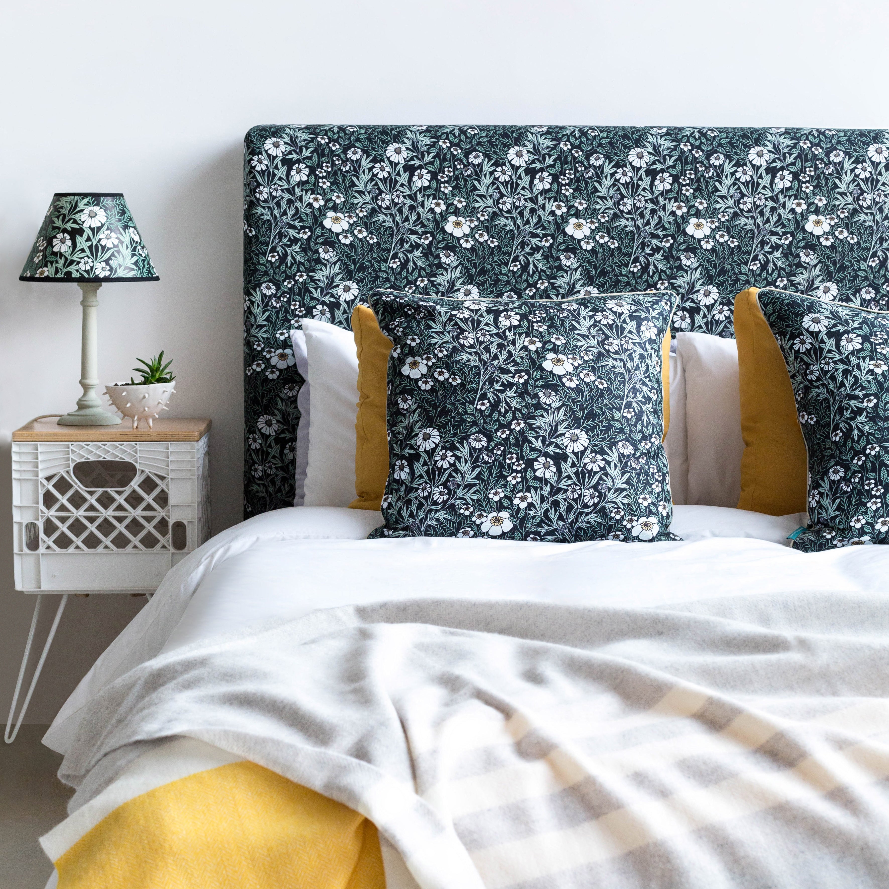 A bed with a headboard and pillows upholstered in a dense floral print in white, tan and green on a black field.