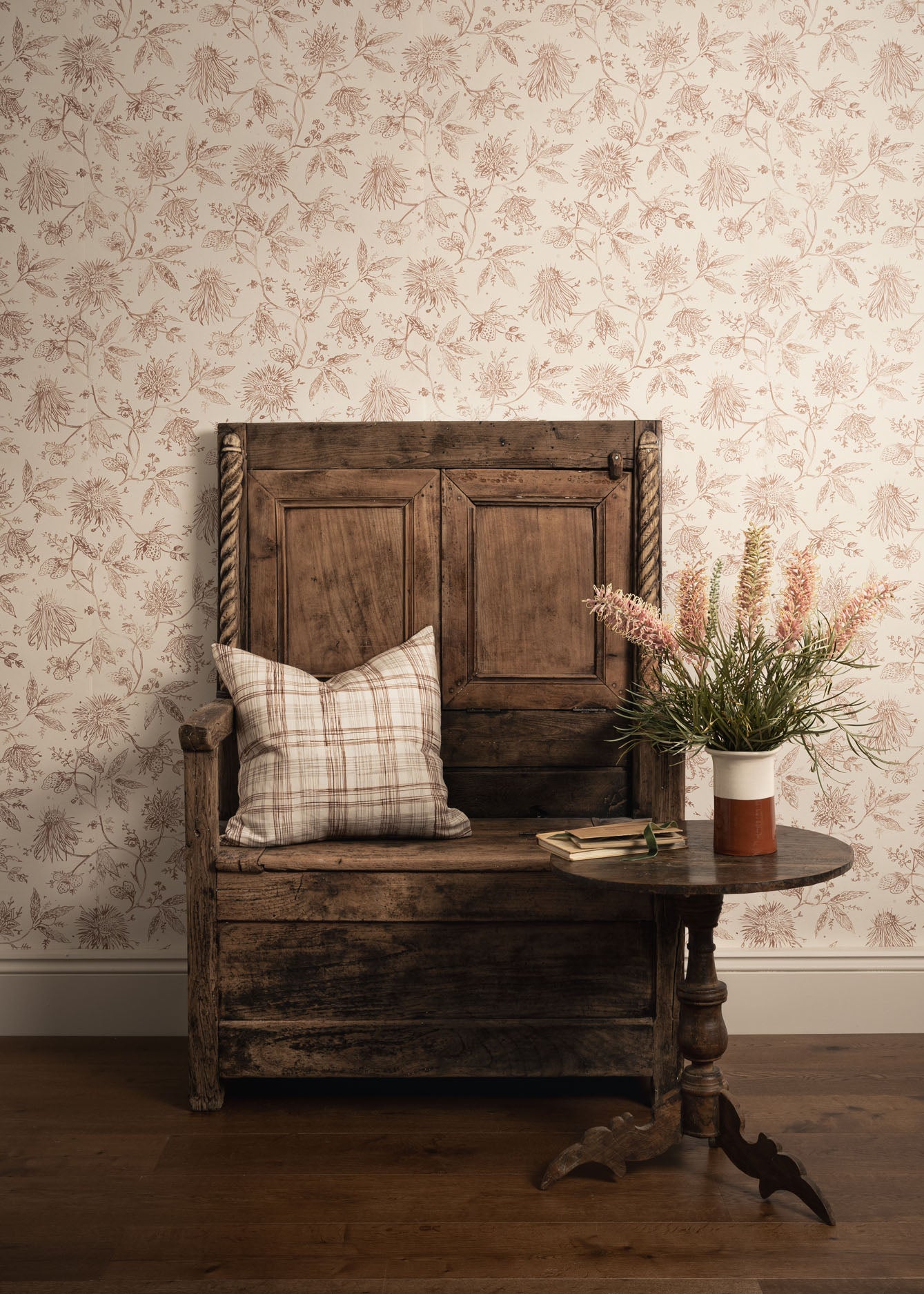 Vintage wooden bench with a plaid pillow and a vase of flowers against a floral wallpapered wall.