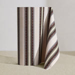 Wallpaper roll in a complex geometric stripe in brown, tan, orange, light purple and black on a cream field.