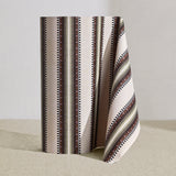 Wallpaper roll in a complex geometric stripe in brown, tan, orange, light purple and black on a cream field.