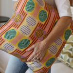 Close up of a woman in white tee shirt and jeans holding a bold geometric patterned pillow in yellow, orange, green and pink. 