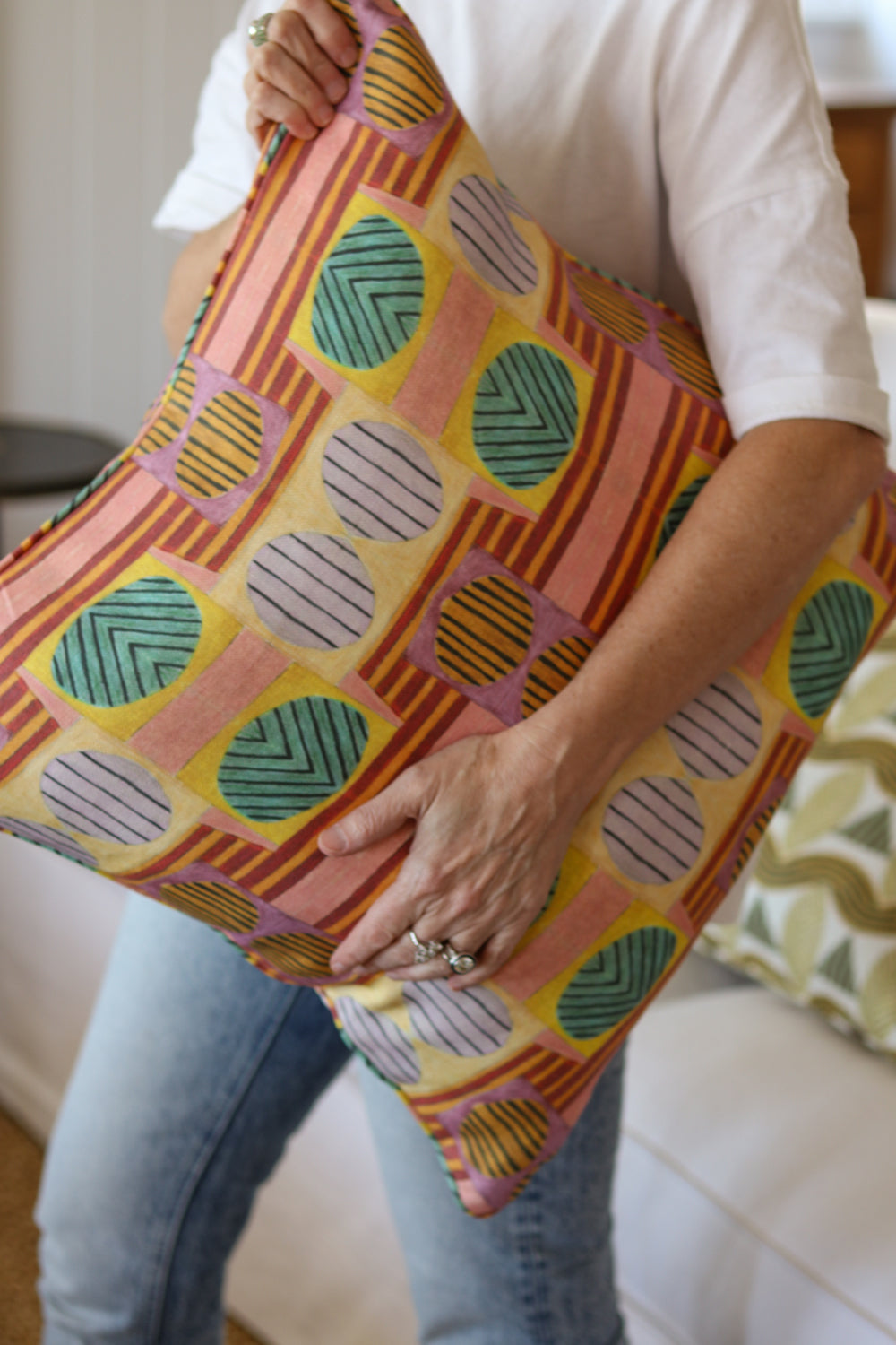 Close up of a woman in white tee shirt and jeans holding a bold geometric patterned pillow in yellow, orange, green and pink. 