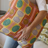 Close up of a woman in white tee shirt and jeans holding a bold geometric patterned pillow in yellow, orange, green and pink. 