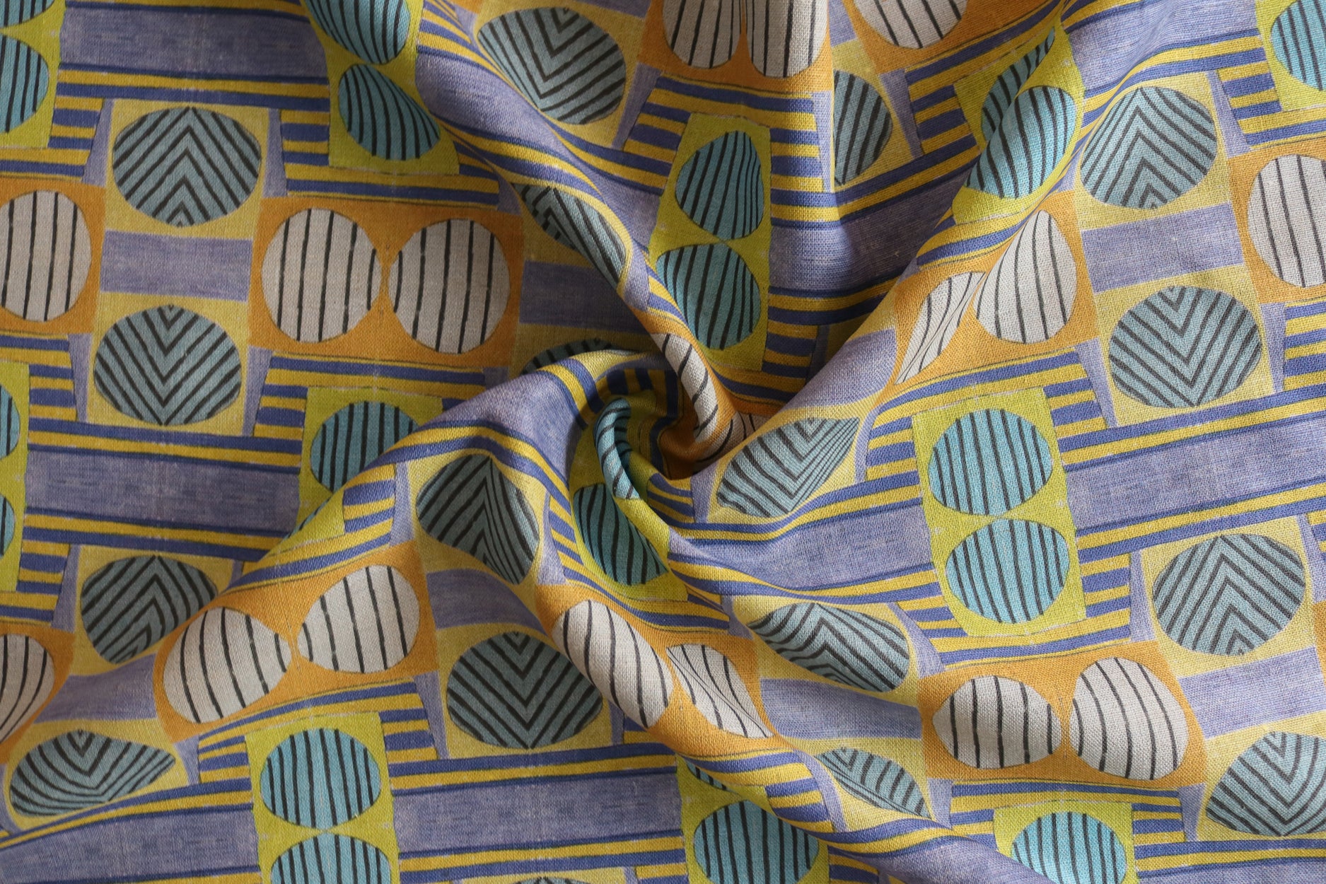 Close up of gently draped printed fabric with a complex geometric and striped pattern with yellow, blue, orange and pale pink. 