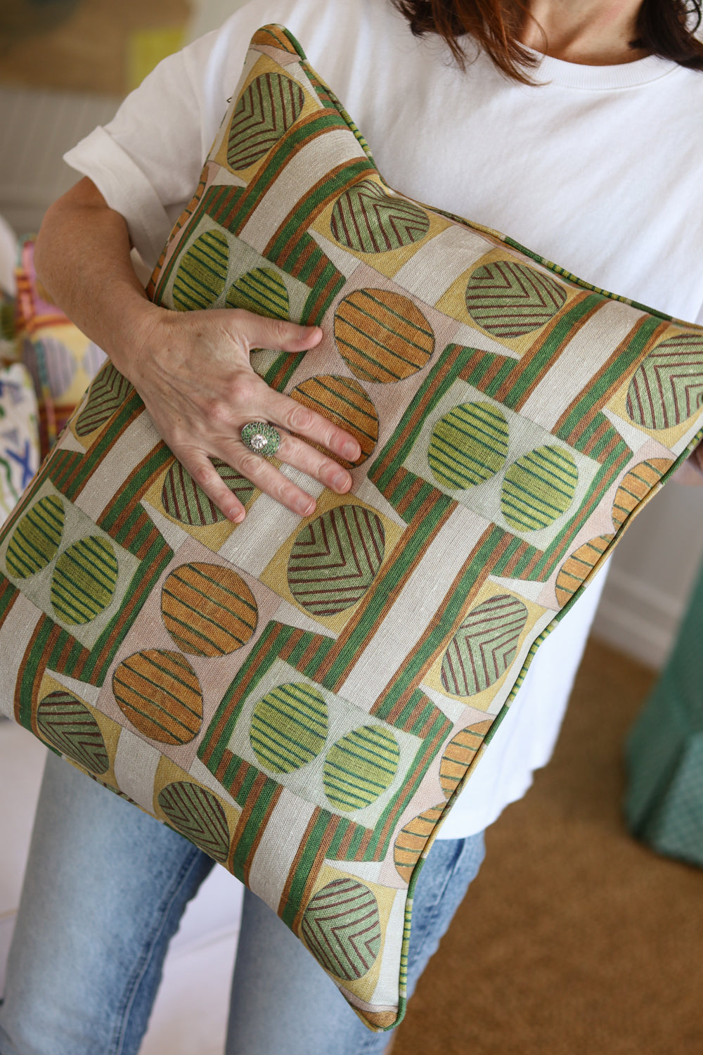Close up of a woman in white tee shirt and jeans holding a bold geometric patterned pillow in orange, brown and green. 