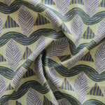 A close-up of a gently draped patterned fabric featuring repeating rows of stylized grey leaves, blue and yellow stripe triangles and wavy blue-yellow lines on yellow background.
