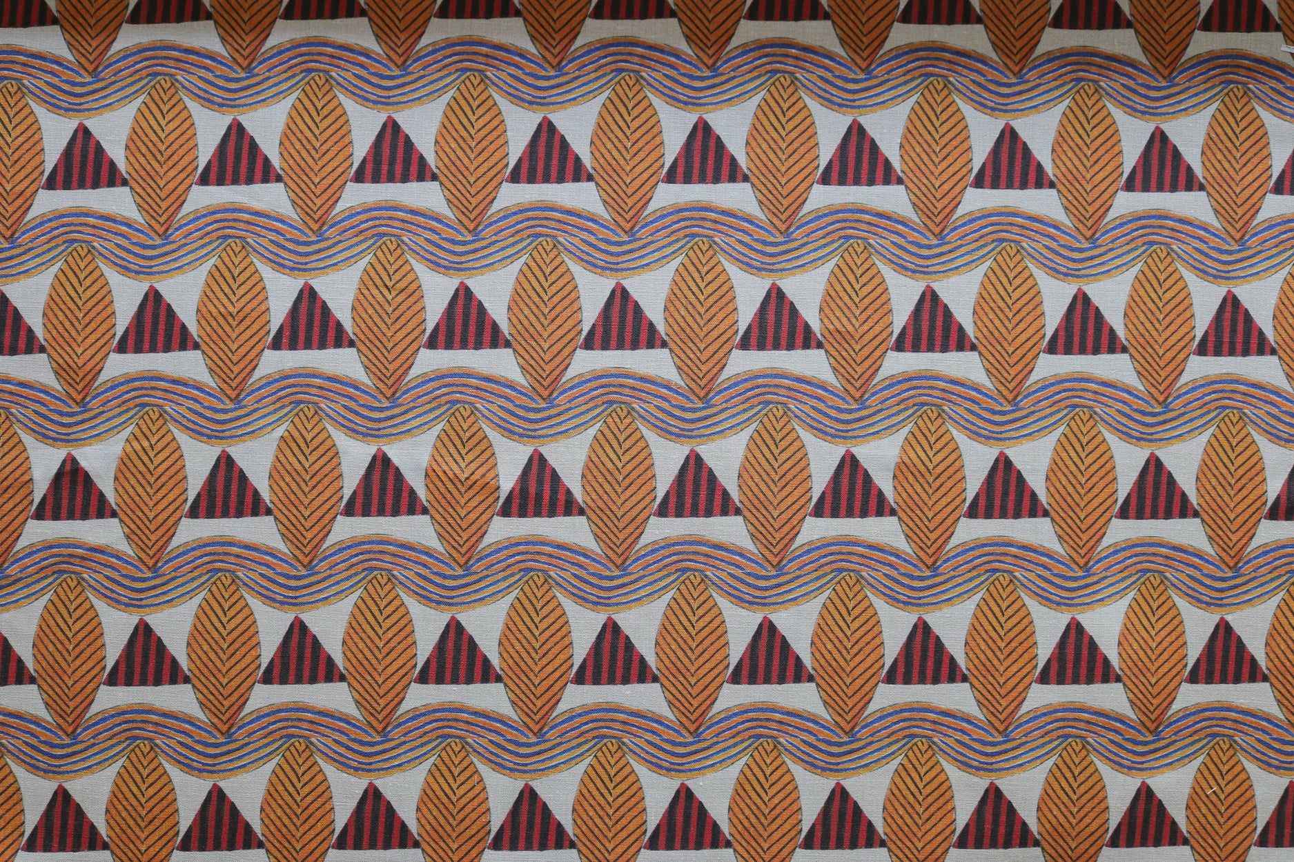 A close-up of a patterned fabric featuring repeating rows of stylized orange leaves, navy and red striped triangles and wavy orange-blue lines on a white background