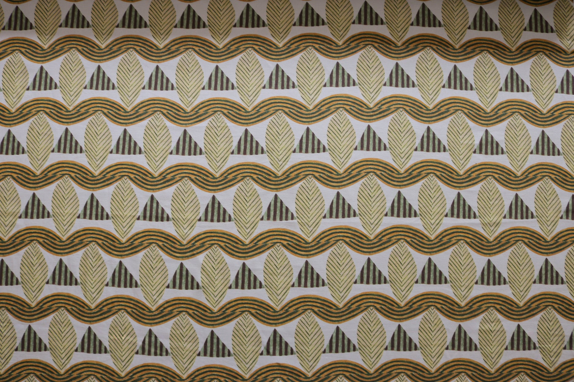 A close-up of a patterned fabric featuring repeating rows of stylized tan leaves, brown and green striped triangles and wavy tan-green lines on a white background