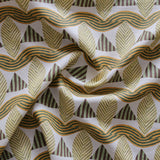 A close-up of a gently draped patterned fabric featuring repeating rows of stylized tan leaves, brown and green striped triangles and wavy tan-green lines on a white background