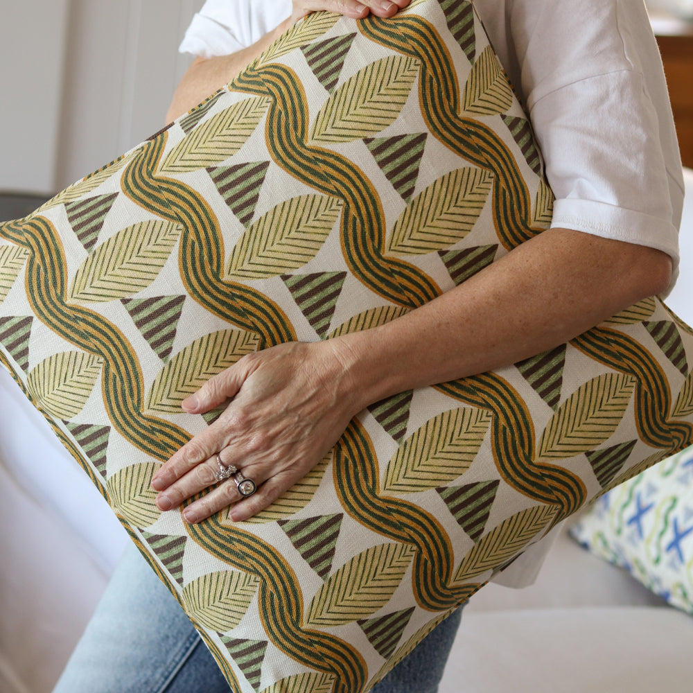 Detail of a woman in a white shirt and jeans holding a bold abstract leaf patterned throw pillow in tan and green in front of a white couch. 