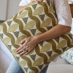 Detail of a woman in a white shirt and jeans holding a bold abstract leaf patterned throw pillow in tan and green in front of a white couch. 
