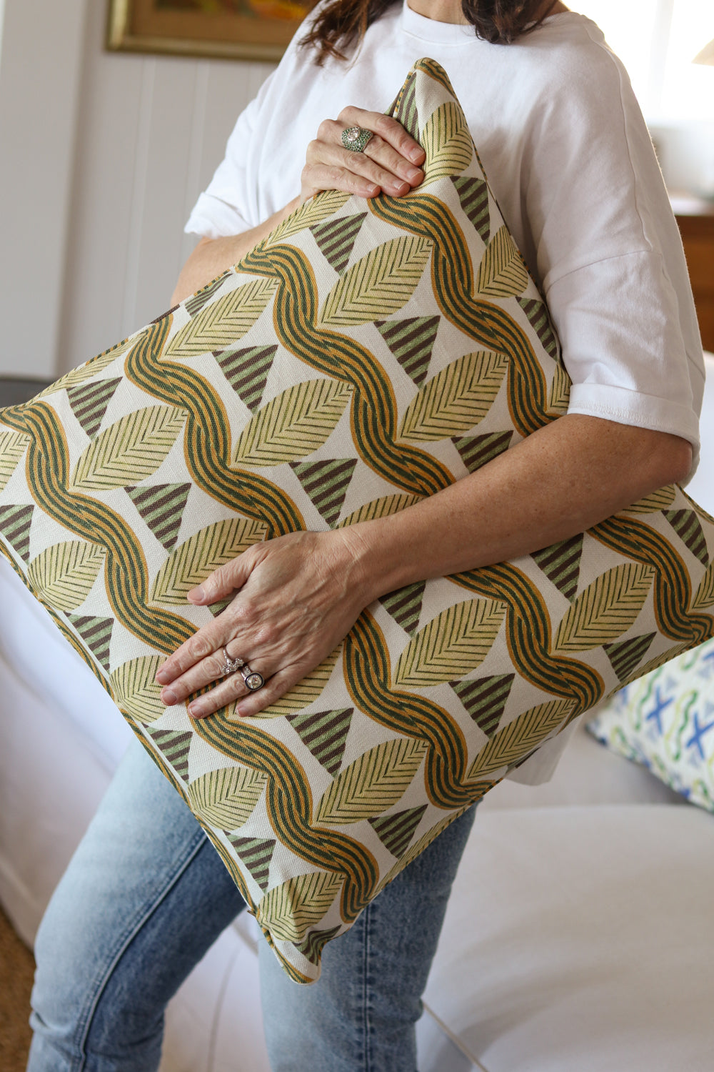 Detail of a woman in a white shirt and jeans holding a bold abstract leaf patterned throw pillow in tan and green in front of a white couch. 