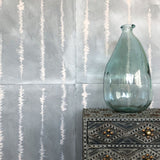 A vase on an end table stands in front of a wall papered in an irregular vibrating linear pattern in peach on a greige field.