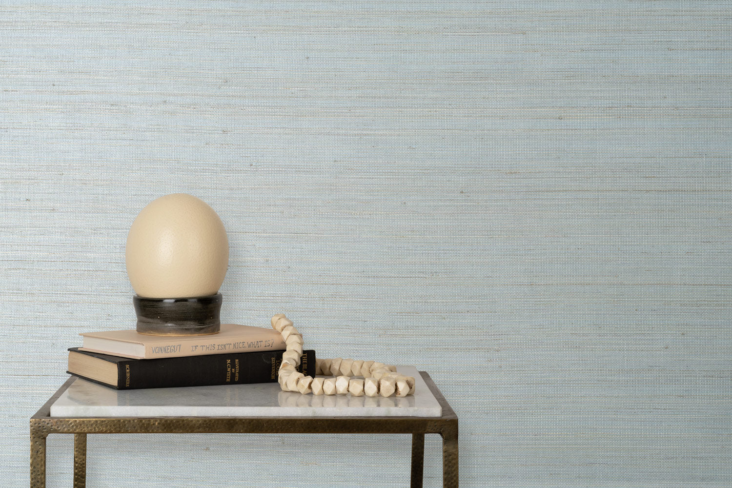 End table with books and a lamp in front of a wall papered in textured grasscloth in shades of pastel, blue and turquoise.