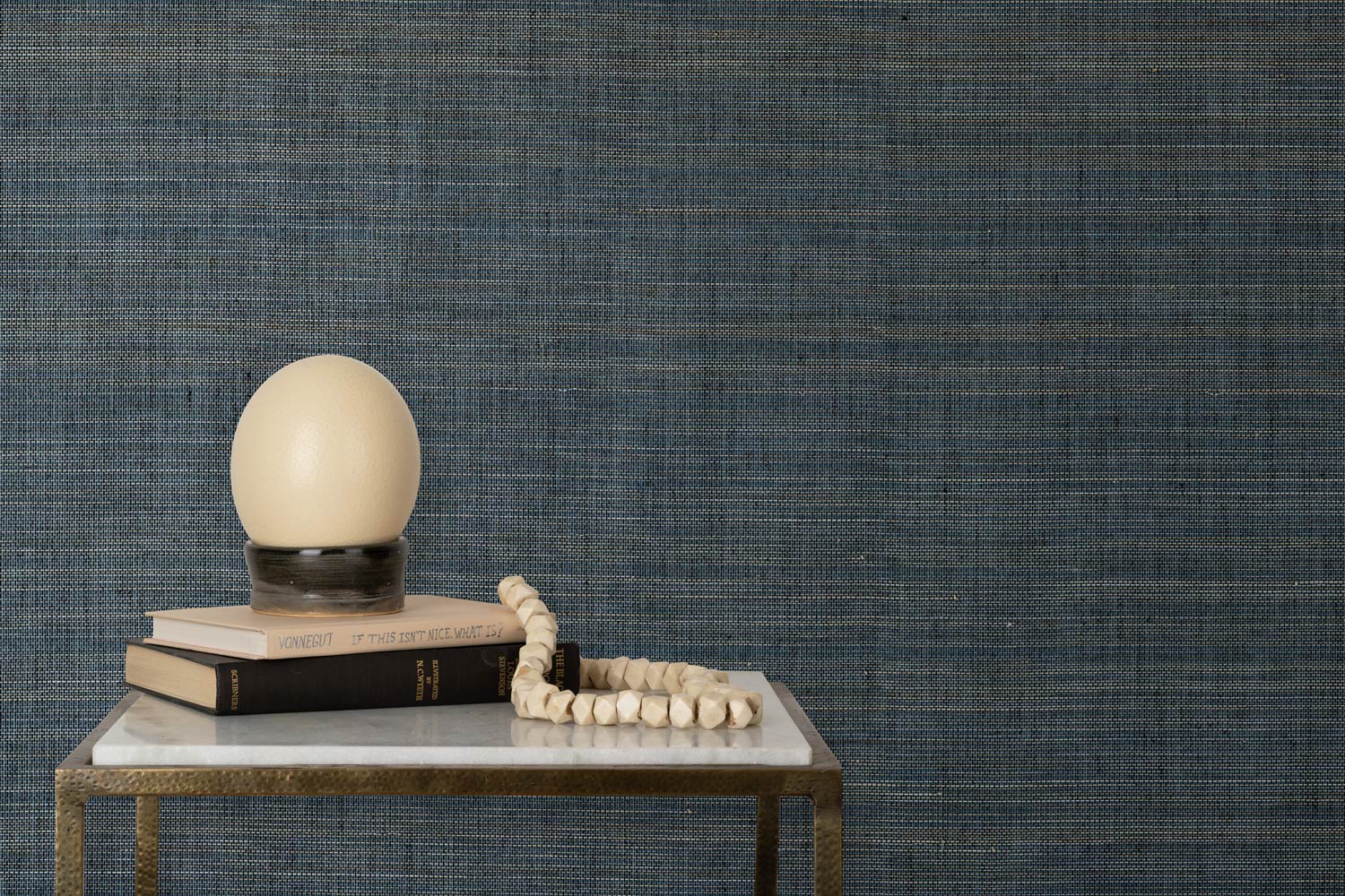 End table with books and a lamp in front of a wall papered in textured grasscloth in shades of green, cream and turquoise.