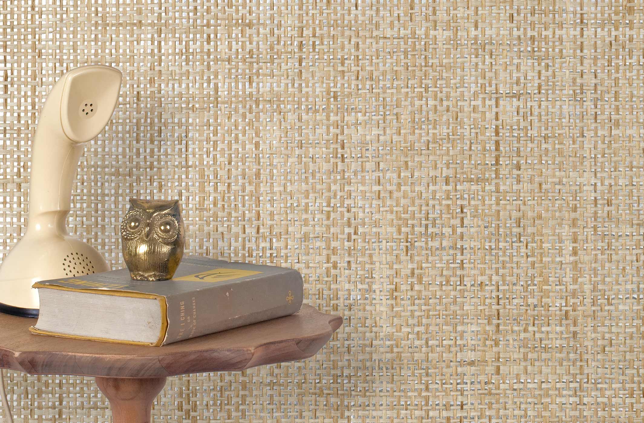 End table with a phone, book and owl sculpture in front of a wall covered in paperweave grasscloth in metallic tan and gray.