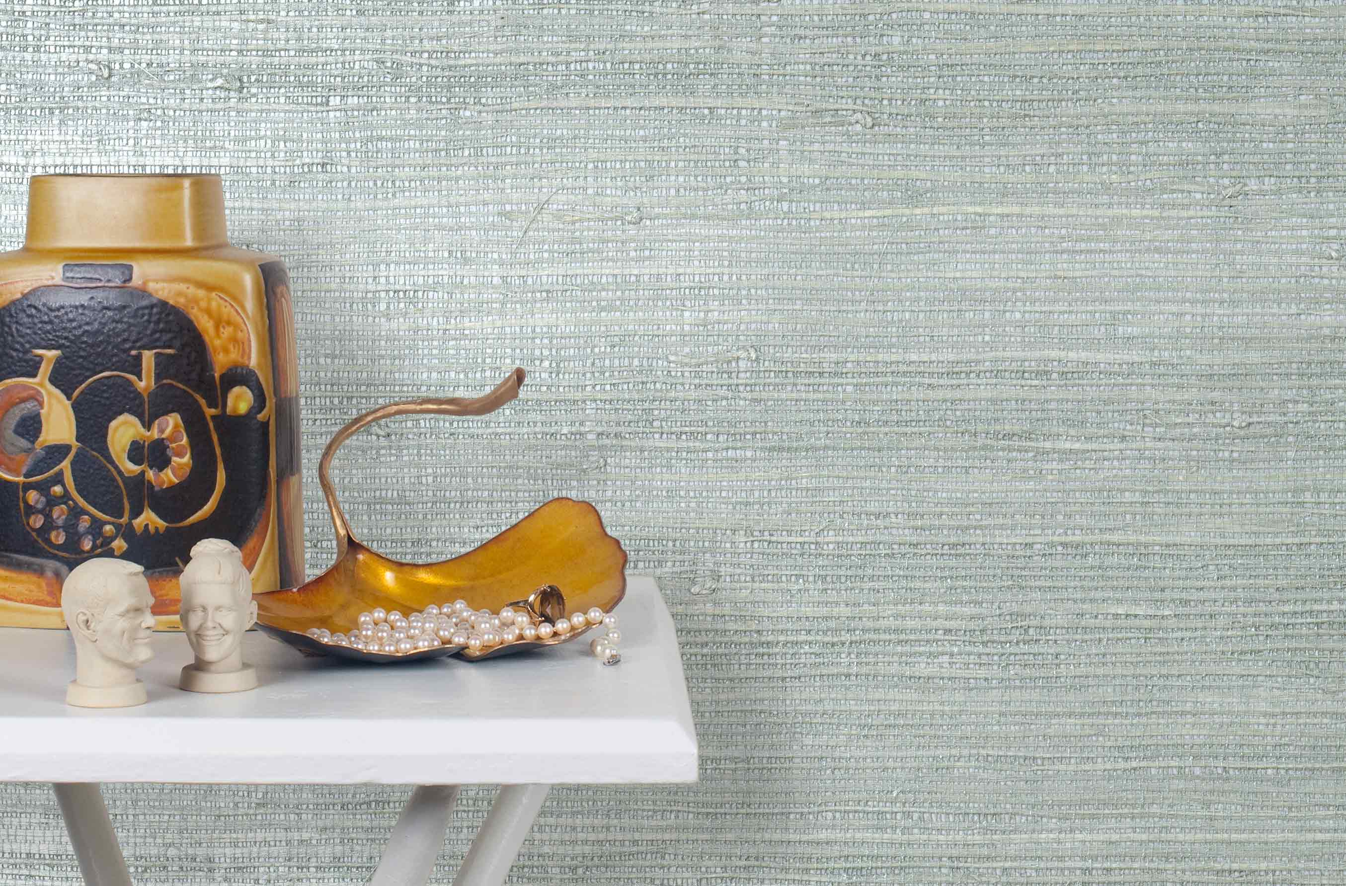 End table with a vase and sculptures in front of a wall papered in metallic-rubbed jute grasscloth in blue pastel.
