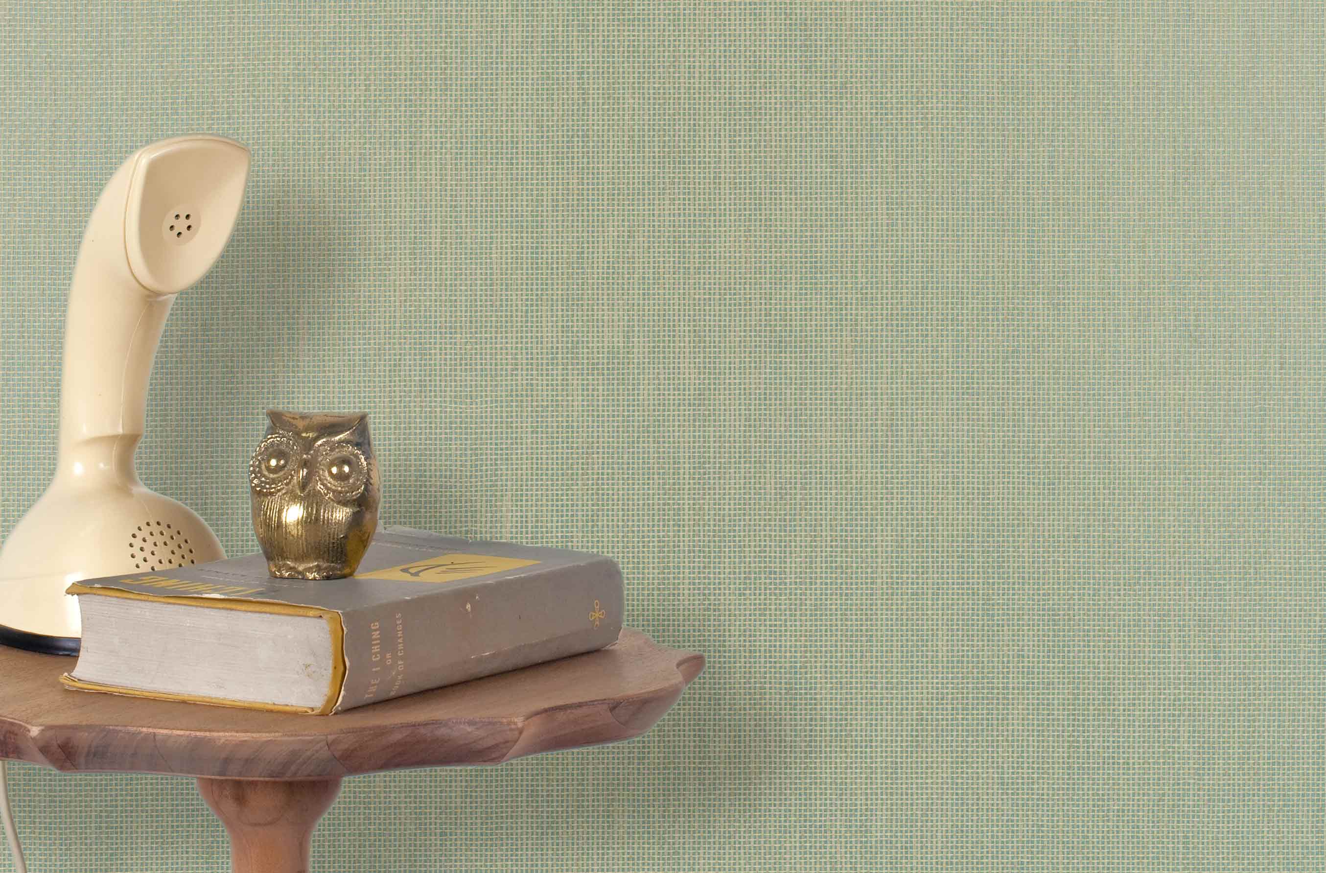 End table with a phone, book and owl sculpture in front of a wall covered in paperweave grasscloth in seafoam green.