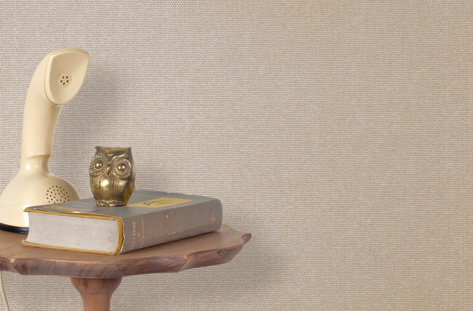 End table with a phone, book and owl sculpture in front of a wall covered in metallic paperweave grasscloth in neutral.
