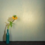 A vase of flowers stands in front of a wall papered in an abstract ridged texture in metallic green.