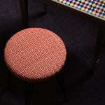 Overhead image of an upholstered stool in a red embroidered fabric with a harlequin enameled side table and stack of books. 