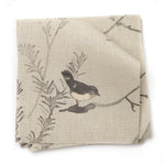 A stack of fabric swatches in a painterly bird and branch pattern in shades of gray on a tan field.