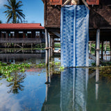 Woman holding a large blue and white patterned fabric in front of a wooden house on stilts on a waterway.
