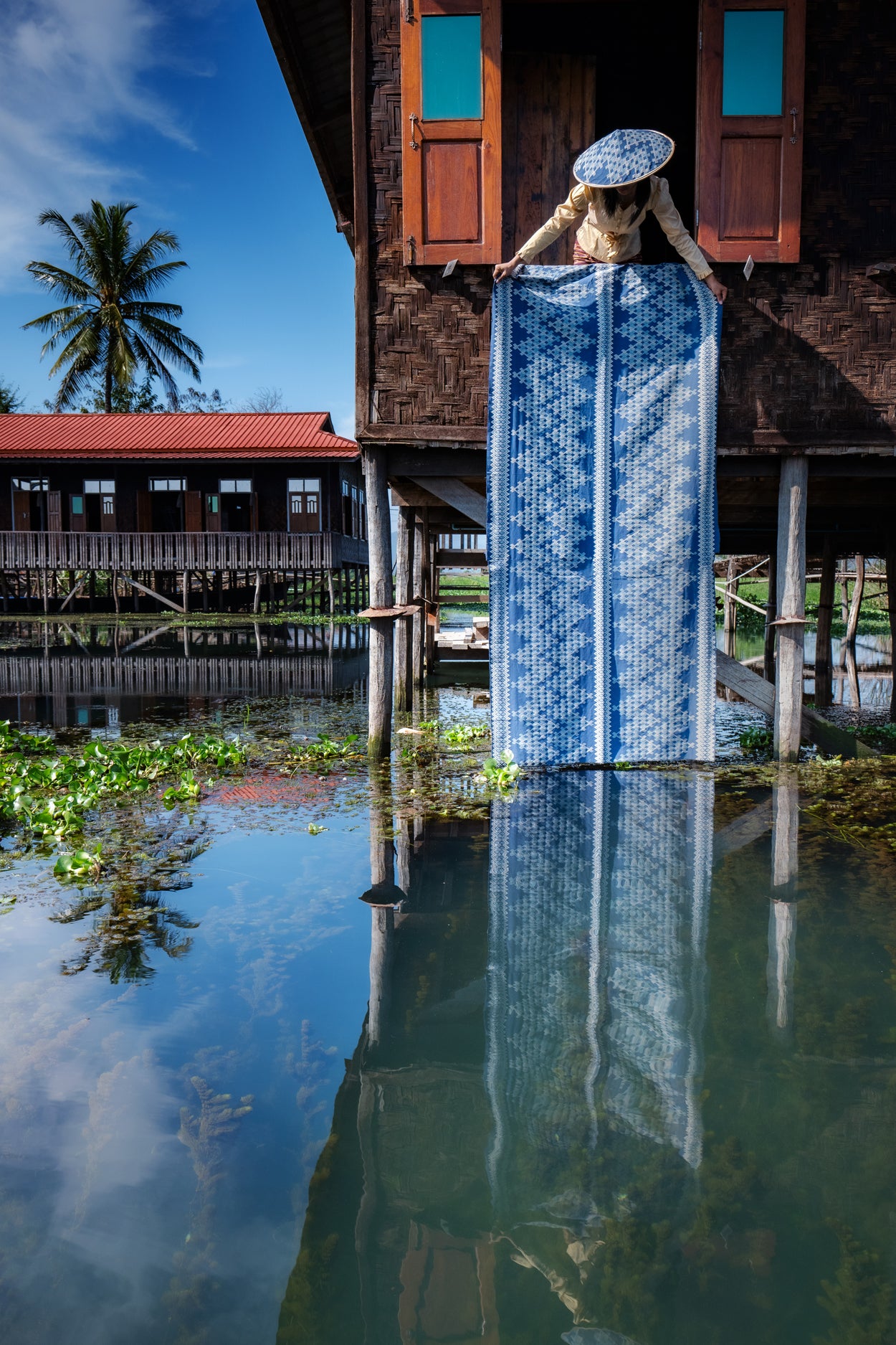 Woman holding a large blue and white patterned fabric in front of a wooden house on stilts on a waterway.